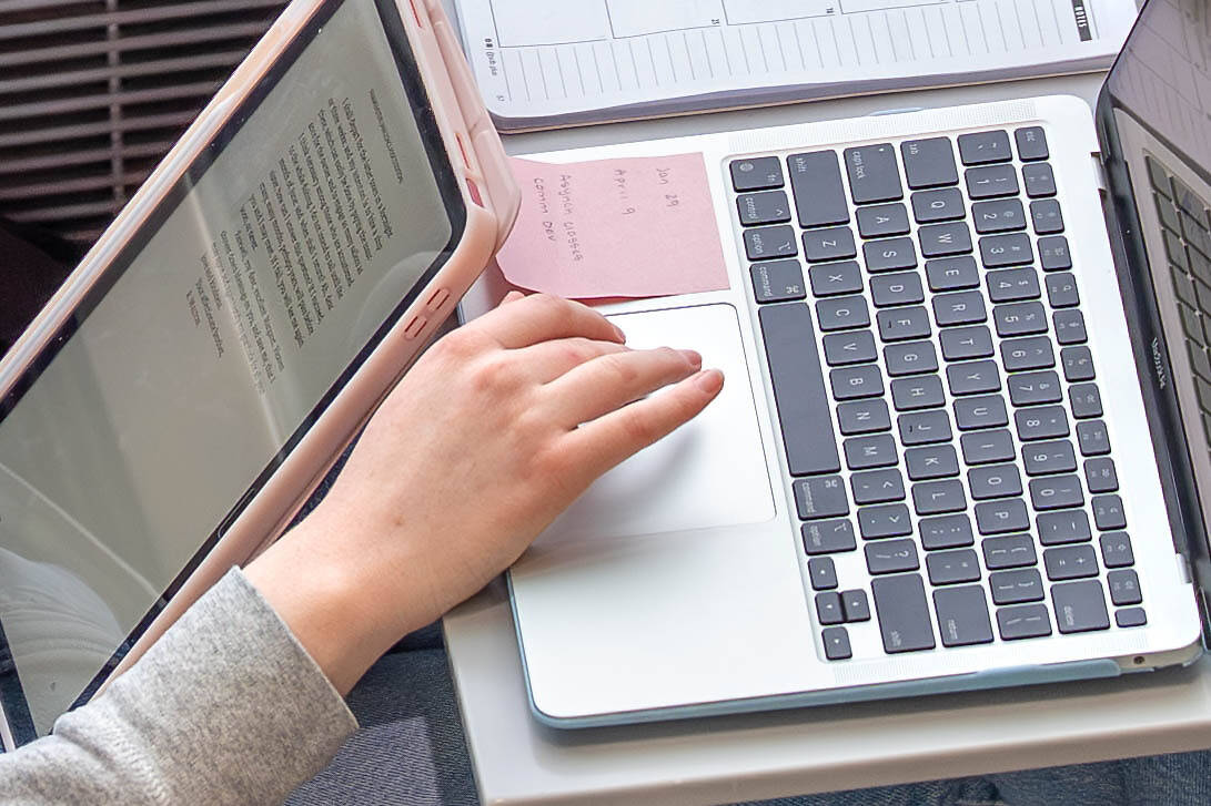 A person's hand rests on a laptop's touchpad while an e-reader displays text nearby.